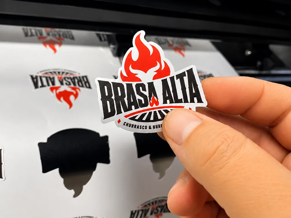 Adesivo já recortado no contorno sendo destacado da folha, na saída da plotter